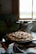 A freshly baked round cake or bread rests in a dark skillet on top of a decorative cloth with floral and geometric patterns. The scene is lit softly from a window in the background, which casts a warm glow over the kitchen area.