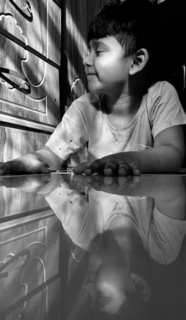 A child with short, dark hair is sitting and smiling softly, their face turned sideways. The child is wearing a light-colored t-shirt, with a reflective surface in front of them showing their reflection. The background features a wooden texture with decorative patterns, and light creates interesting shadows on the wall.
