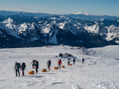 A group of people equipped with hiking gear are navigating a snow-covered mountain landscape. Some are pulling orange sleds, possibly carrying supplies. The expansive scene includes rugged snowy peaks and a distant range of mountains under a clear blue sky.
