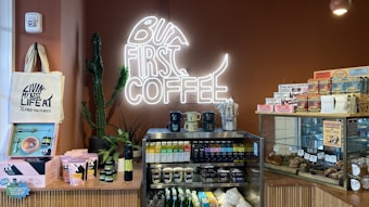 A coffee shop counter displaying various products, including mugs, tea tins, packaged coffee, and pastries. A neon sign with 'But First Coffee' is mounted on a brown wall. A tote bag and several plants add decoration to the scene.