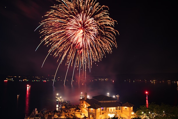 Bright, colorful fireworks burst in the night sky over a large body of water, with reflections visible on the surface. Below, a well-lit building and an array of boats are visible, indicating a festive or celebratory atmosphere.