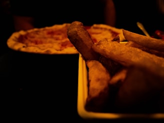 A close-up of a serving of thick-cut fries in a tray with a pizza in the background, both set in a dimly lit setting.