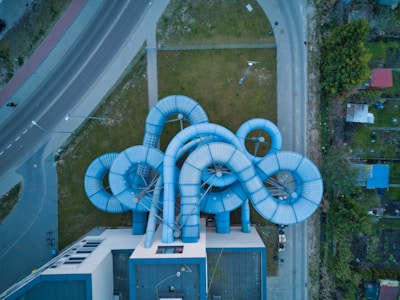 Aerial view of a series of interconnected blue water slide tubes attached to the side of a building. The structure sprawls out over a lawn, surrounded by roads and a few trees. The slide loops and spirals in multiple directions, creating a complex and intricate pattern.