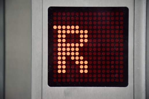 A digital display panel features a large orange letter 'R' made up of illuminated dots against a dark background. The panel is bordered by a metallic frame.