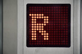A digital display panel features a large orange letter 'R' made up of illuminated dots against a dark background. The panel is bordered by a metallic frame.