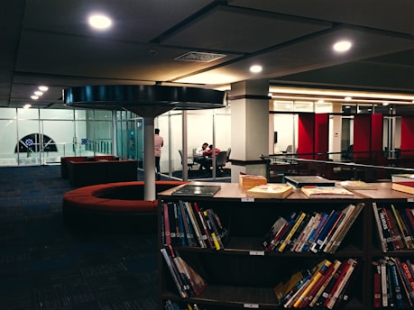 A library scene with bookshelves filled with books in the foreground. There is a seating area with circular red couches and tables. In the background, a glass-walled room is visible with people working at a table. The lighting is warm and the space appears modern.