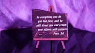 A small easel holds a rectangular plaque with an inspirational quote from Proverbs 3:6, written in white lettering on a dark background. The setup is bathed in purple lighting, giving the scene a warm, contemplative atmosphere.
