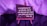 A small easel holds a rectangular plaque with an inspirational quote from Proverbs 3:6, written in white lettering on a dark background. The setup is bathed in purple lighting, giving the scene a warm, contemplative atmosphere.