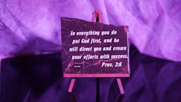 A small easel holds a rectangular plaque with an inspirational quote from Proverbs 3:6, written in white lettering on a dark background. The setup is bathed in purple lighting, giving the scene a warm, contemplative atmosphere.