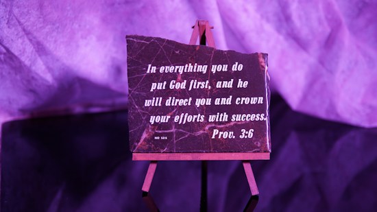 A small easel holds a rectangular plaque with an inspirational quote from Proverbs 3:6, written in white lettering on a dark background. The setup is bathed in purple lighting, giving the scene a warm, contemplative atmosphere.