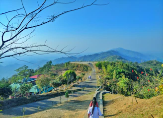 woman in white shirt walking on pathway near green trees and mountain during daytime