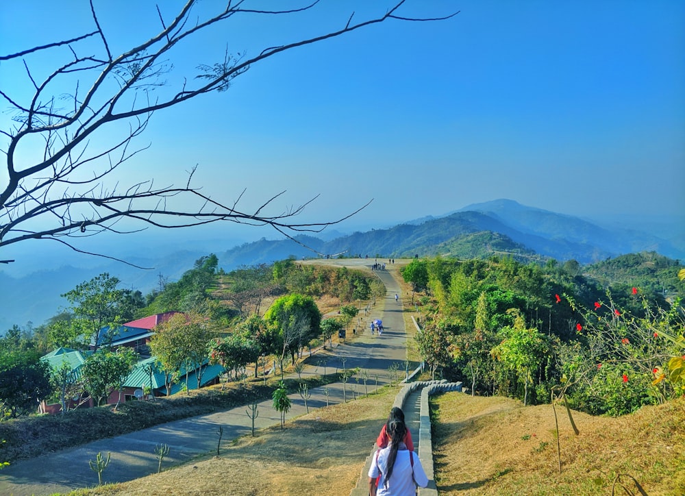 woman in white shirt walking on pathway near green trees and mountain during daytime