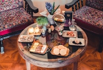 A round wooden table is set with a traditional aesthetic, featuring various cultural and culinary items. There are decorative bowls, plates with snacks, fruits, and a small vase with a blue flower arrangement. In the background, two ornate chairs with vibrant, patterned fabrics complement the warm, cozy ambiance.
