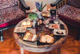 A round wooden table is set with a traditional aesthetic, featuring various cultural and culinary items. There are decorative bowls, plates with snacks, fruits, and a small vase with a blue flower arrangement. In the background, two ornate chairs with vibrant, patterned fabrics complement the warm, cozy ambiance.