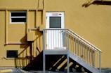 A yellow building exterior with a white door elevated by a metal staircase. The door has signs above it and the stairway landing. The building's surface features pipes, utility boxes, and a partially visible window.
