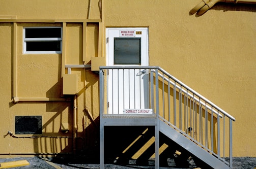 A yellow building exterior with a white door elevated by a metal staircase. The door has signs above it and the stairway landing. The building's surface features pipes, utility boxes, and a partially visible window.