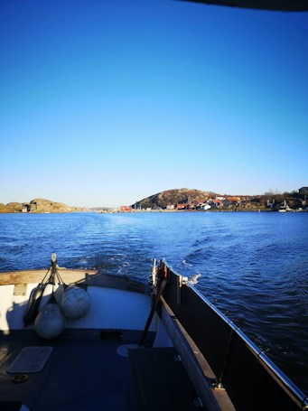 A small boat traverses calm blue waters, with a view towards a coastal landscape featuring small hills and a cluster of colorful buildings. The sun casts a clear light across the scene, creating vivid reflections on the water. In the foreground, the deck of the boat is visible, with some mooring equipment attached.
