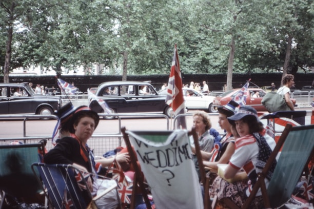 A group of people are sitting on deck chairs decorated with union jack flags, on a street lined with trees and metal barriers. Some are wearing hats with patriotic designs. There are classic cars and people walking in the background.