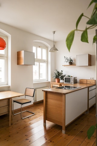 A bright and modern kitchen interior featuring wooden flooring and minimalistic design. In the center, there's an island counter with a built-in stovetop. The kitchen walls have white cabinetry and shelves with potted plants. A dining area with a wooden table and a black mesh chair is visible. Natural light streams in through large arched windows.
