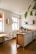A bright and modern kitchen interior featuring wooden flooring and minimalistic design. In the center, there's an island counter with a built-in stovetop. The kitchen walls have white cabinetry and shelves with potted plants. A dining area with a wooden table and a black mesh chair is visible. Natural light streams in through large arched windows.
