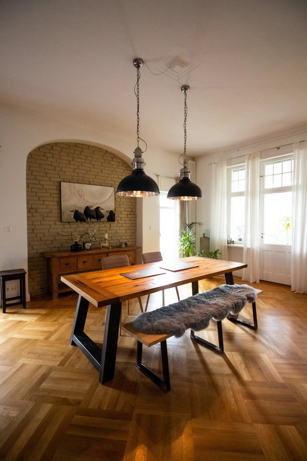 A finished wooden dining table and bench in a bright dining room under two pendant lamps, a sideboard against the brick wall behind.