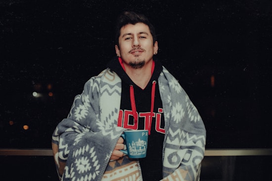 A person is wrapped in a patterned blanket while holding a blue mug. The person is wearing a black hoodie with red and white lettering and has a relaxed expression. The background is dark with some blurred lights.