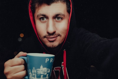 A person wearing a red-hooded sweatshirt holds a blue mug with white designs and the word 'PR' on it. The background is dark, with small blurred lights visible. The person appears to be smiling softly at the camera.