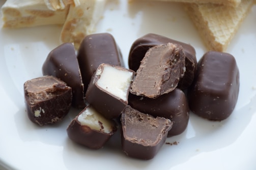A collection of chocolate-covered confectionery pieces scattered across a white surface. Several of the chocolates have been cut open, revealing a creamy interior. In the background, there are light-colored wafer biscuits.