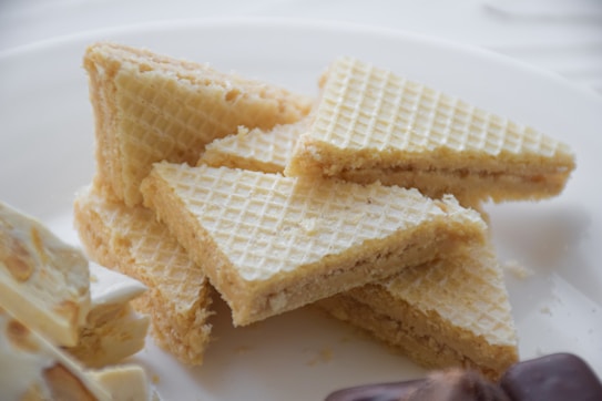 A pile of triangular wafer cookies with light patterns on a white plate. The wafers have a creamy filling and a lightly textured surface. Beside them, there are pieces of a white chocolate bar with nuts.
