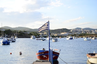 Several boats are anchored in a calm harbor, with a Greek flag prominently displayed on a pole in the foreground. The background features rolling hills and a cityscape under a partly cloudy sky.