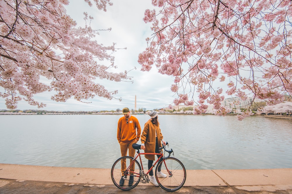 man in yellow jacket and black pants standing beside black bicycle near body of water during