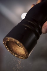 A close-up view of a pepper grinder with ground pepper particles falling from it. The grinder is being held at an angle, highlighting its dark color and metallic base where the pepper disperses.