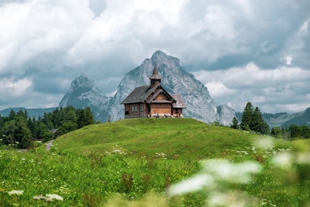 brown wooden house on green grass field near mountain under white clouds during daytime