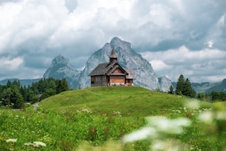 brown wooden house on green grass field near mountain under white clouds during daytime