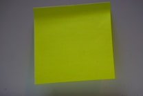 A bright yellow sticky note positioned against a plain background, slightly shadowed around the edges due to lighting.
