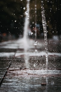 Jets of water shoot up from a fountain, creating droplets that glisten in the air. The scene is captured in a shallow depth of field, focusing on the central stream of water while the background remains blurred. The pavement is wet, reflecting the splashes of the fountain.