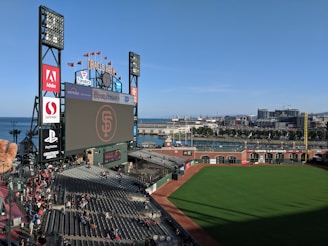 A baseball stadium with a large screen displaying a logo in the center. The stands are partially filled with spectators, and there are various advertisements surrounding the screen. The view includes a green field, with buildings and water visible in the background, suggesting the stadium is near a waterfront city area.