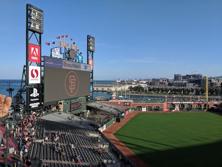 A baseball stadium with a large screen displaying a logo in the center. The stands are partially filled with spectators, and there are various advertisements surrounding the screen. The view includes a green field, with buildings and water visible in the background, suggesting the stadium is near a waterfront city area.