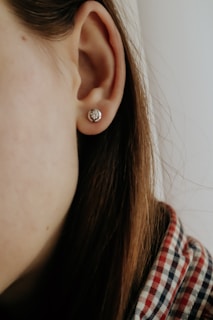 A close-up of a person's ear adorned with a small, round earring. The person's hair is brown and partially visible, and they are wearing a plaid shirt with red and blue checks. The background is soft and out of focus.