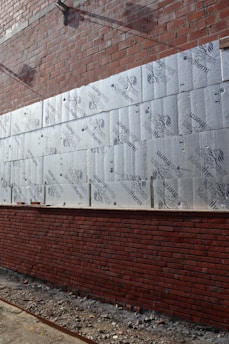 A partially constructed brick wall with layers of insulation boards attached to it. The insulation boards are marked with the brand 'Kingspan Kooltherm.' The bottom half of the wall is complete, while the top half is still under construction. The ground is covered in debris from the construction process.
