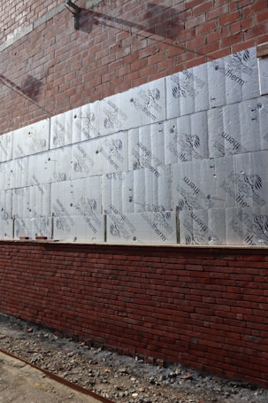 A partially constructed brick wall with layers of insulation boards attached to it. The insulation boards are marked with the brand 'Kingspan Kooltherm.' The bottom half of the wall is complete, while the top half is still under construction. The ground is covered in debris from the construction process.