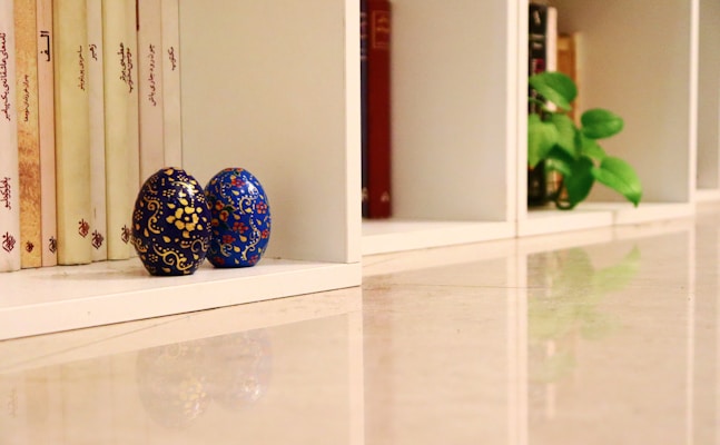 A neatly organized bookshelf holds books with vertical script on their spines. Two intricately designed eggs with blue and black backgrounds and gold patterns rest against the books. A nearby cubby features a small green plant, adding a touch of nature to the serene setting. The polished floor reflects the items, enhancing the tranquil and tidy atmosphere.