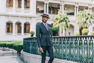 A person wearing a stylish suit and a hat with a colorful band is standing by an ornate metal railing. Behind them, a grand building with tall columns and wooden windows is visible. The area is surrounded by neatly trimmed greenery and palm trees, creating an elegant setting.