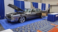 A classic car with an open hood is displayed at an indoor exhibition booth, featuring banners and a table nearby. The vehicle has a shiny exterior with large chrome wheels and text promoting vehicle safety on its side. The setting includes blue and white draped curtains and a carpeted floor with a geometric pattern.