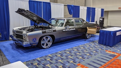 A classic car with an open hood is displayed at an indoor exhibition booth, featuring banners and a table nearby. The vehicle has a shiny exterior with large chrome wheels and text promoting vehicle safety on its side. The setting includes blue and white draped curtains and a carpeted floor with a geometric pattern.