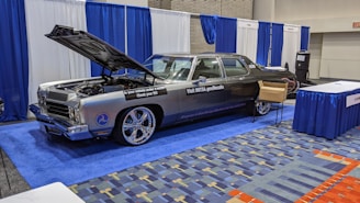A classic car with an open hood is displayed at an indoor exhibition booth, featuring banners and a table nearby. The vehicle has a shiny exterior with large chrome wheels and text promoting vehicle safety on its side. The setting includes blue and white draped curtains and a carpeted floor with a geometric pattern.