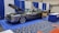 A classic car with an open hood is displayed at an indoor exhibition booth, featuring banners and a table nearby. The vehicle has a shiny exterior with large chrome wheels and text promoting vehicle safety on its side. The setting includes blue and white draped curtains and a carpeted floor with a geometric pattern.
