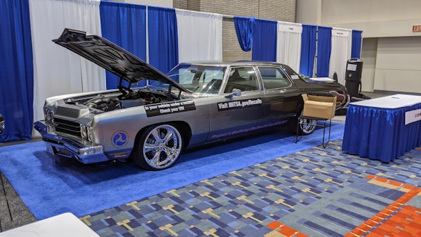 A classic car with an open hood is displayed at an indoor exhibition booth, featuring banners and a table nearby. The vehicle has a shiny exterior with large chrome wheels and text promoting vehicle safety on its side. The setting includes blue and white draped curtains and a carpeted floor with a geometric pattern.