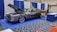 A classic car with an open hood is displayed at an indoor exhibition booth, featuring banners and a table nearby. The vehicle has a shiny exterior with large chrome wheels and text promoting vehicle safety on its side. The setting includes blue and white draped curtains and a carpeted floor with a geometric pattern.