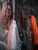 Several fishing nets in various colors including orange, red, and white are hanging against a rough stone wall. Some nets have floats attached, and ropes are tangled among them.